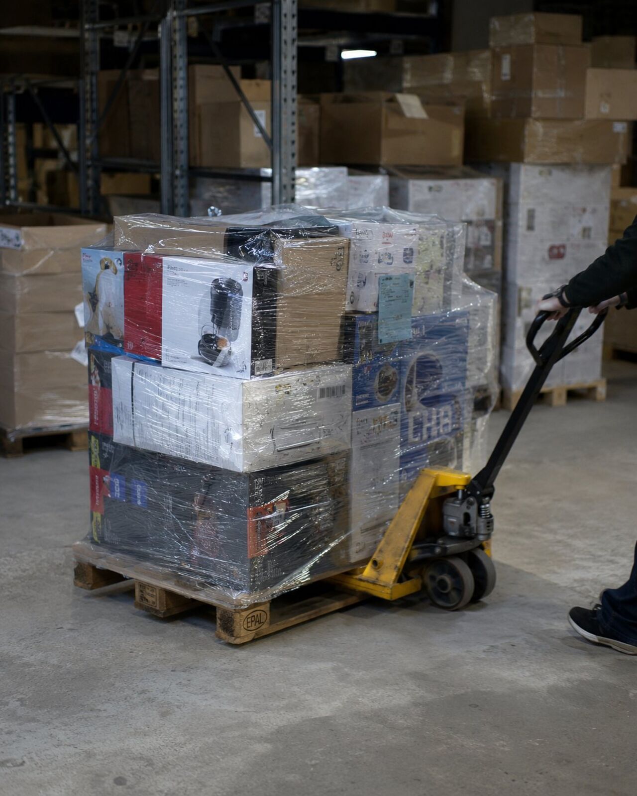A pallet jack moving a shrink-wrapped pallet of mixed branded goods across a warehouse floor